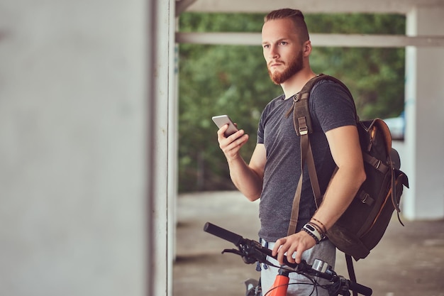 Guapo hombre pelirrojo con un elegante corte de pelo y barba vestido con ropa deportiva con una bicicleta y una mochila, usando el teléfono inteligente parado afuera.