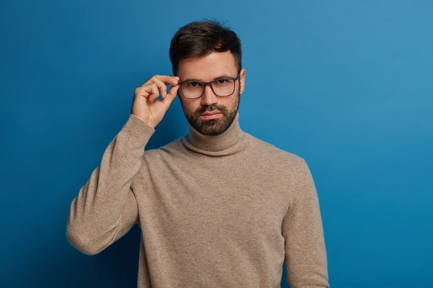 Guapo hombre seguro de sí mismo con cerdas gruesas, mantiene la mano en el borde de las gafas