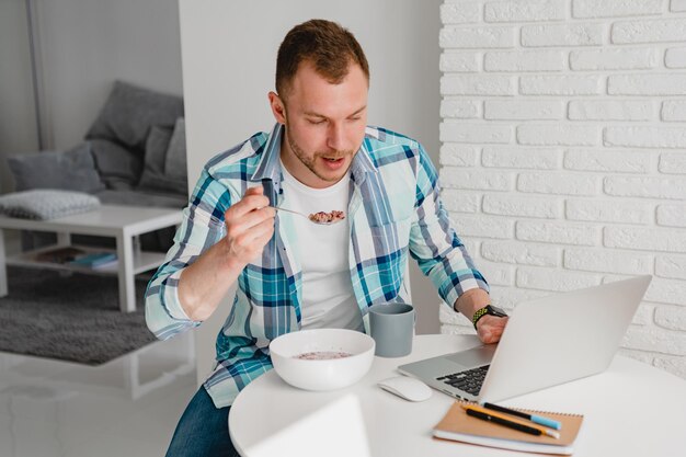 Guapo hombre sonriente en camisa sentado en la cocina en casa en la mesa trabajando en línea en la computadora portátil