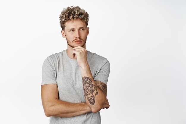 Guapo hombre tatuado rubio mirando hacia otro lado con cara pensativa pensando de pie en camiseta gris sobre fondo blanco.
