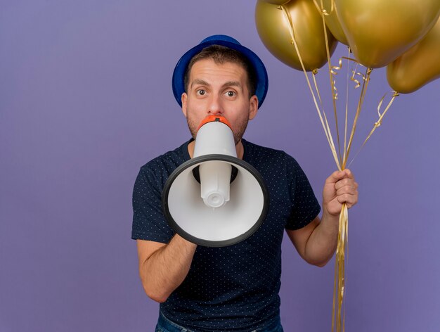 Guapo impresionado con sombrero azul sostiene globos de helio y habla por el altavoz aislado en la pared púrpura