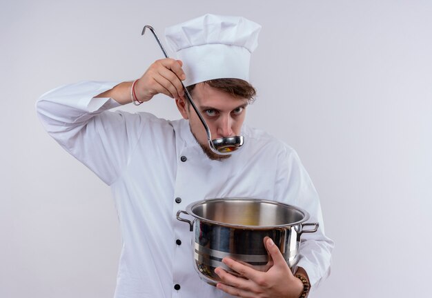 Un guapo joven chef barbudo vestido con uniforme de cocina blanco y sombrero bebiendo sopa de una cacerola con cucharón mientras mira en una pared blanca
