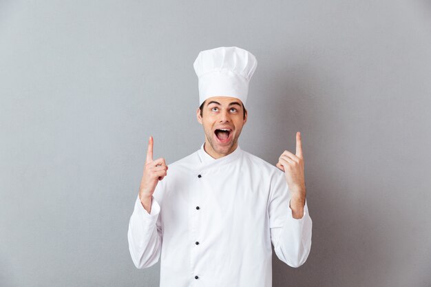 Guapo joven cocinero en uniforme apuntando.