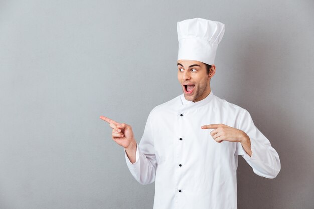 Guapo joven cocinero en uniforme apuntando.