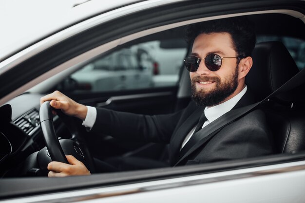 Guapo joven conductor barbudo sonriente en traje completo con cinturón de seguridad de sujeción conduciendo un coche blanco nuevo