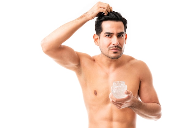 Guapo joven modelo masculino aplicando gel de peinado en su cabello sobre fondo blanco.