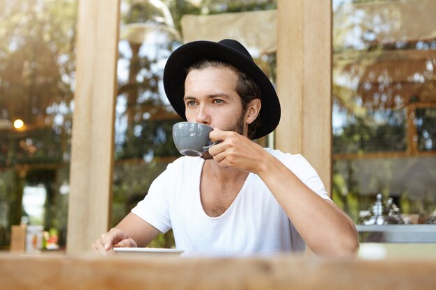 Guapo joven varón caucásico en sombrero sosteniendo una taza de café