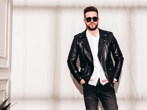 Guapo modelo confiado Hombre elegante sexy en chaqueta de cuero de motociclista y jeans negros Hombre hipster de moda posando junto a la pared blanca en el interior del estudio en el día soleado Sombra desde la ventana