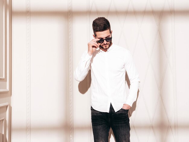 Guapo modelo confiado Hombre elegante sexy vestido con camisa blanca y jeans Hombre hipster de moda posando junto a la pared blanca en el interior del estudio en el día soleado Sombra desde la ventana En gafas de sol