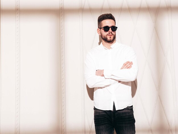 Guapo modelo confiado Hombre elegante sexy vestido con camisa blanca y jeans Hombre hipster de moda posando junto a la pared blanca en el interior del estudio en el día soleado Sombra desde la ventana En gafas de sol