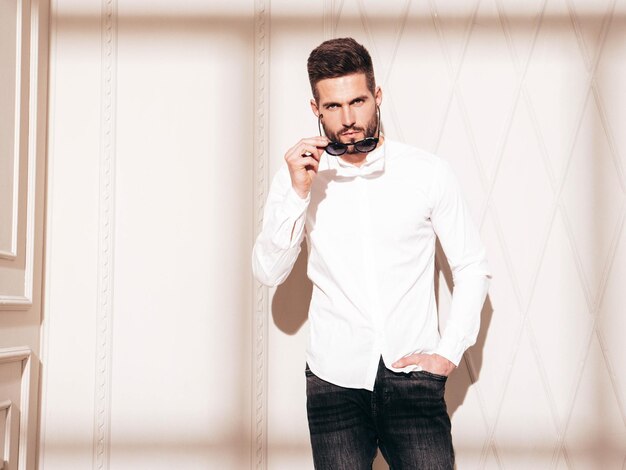 Guapo modelo confiado Hombre elegante sexy vestido con camisa blanca y jeans Hombre hipster de moda posando junto a la pared blanca en el interior del estudio en el día soleado Sombra desde la ventana Quitándose las gafas de sol