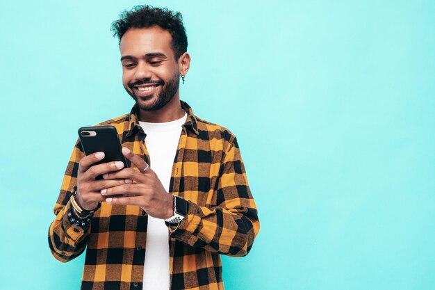 Guapo modelo hipster sonriente Hombre sexy sin afeitar vestido con camisa amarilla de verano y ropa de jeans Hombre de moda posando cerca de la pared azul Sosteniendo teléfono inteligente Mirando la pantalla del teléfono celular Usando aplicaciones