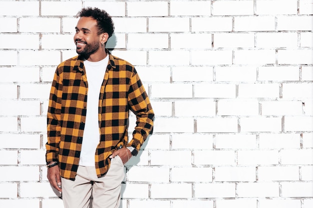 Foto gratuita guapo modelo hipster sonriente hombre sexy sin afeitar vestido con camisa amarilla de verano y ropa de jeans hombre de moda posando junto a la pared de ladrillo blanco en el estudio en un día soleado