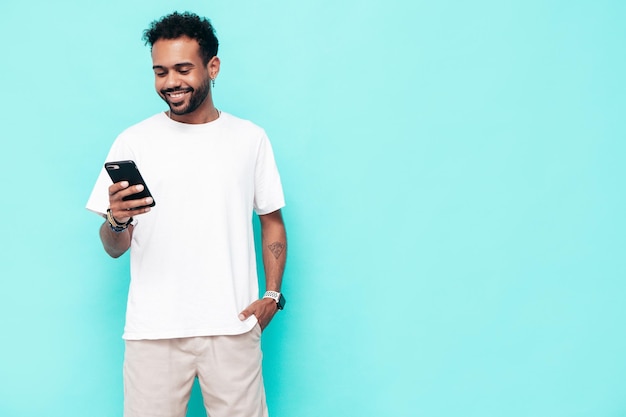 Guapo modelo hipster sonriente Hombre sexy sin afeitar vestido con camiseta blanca de verano y ropa de jeans Hombre de moda posando cerca de la pared azul Sosteniendo el teléfono inteligente Mirando la pantalla del teléfono celular Usando aplicaciones