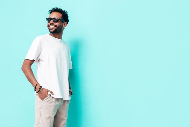 Guapo modelo hipster sonriente Hombre sexy sin afeitar vestido con camiseta blanca de verano y ropa de jeans Hombre de moda posando junto a la pared azul en el estudio Aislado en gafas de sol