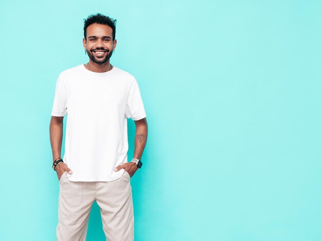 Guapo modelo hipster sonriente Hombre sexy sin afeitar vestido con camiseta blanca de verano y ropa de jeans Hombre de moda posando junto a la pared azul en el estudio Aislado