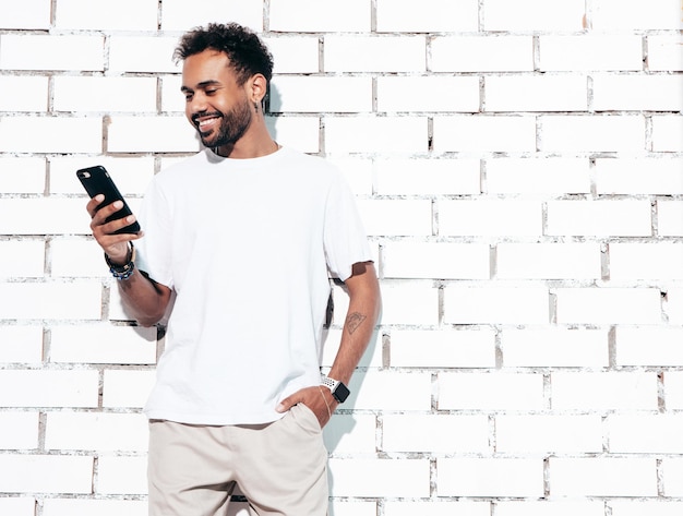 Guapo modelo hipster sonriente Hombre sexy sin afeitar vestido con camiseta blanca de verano y ropa de jeans Hombre de moda posando junto a la pared de ladrillo blanco Mirando la pantalla del teléfono inteligente Usando aplicaciones telefónicas