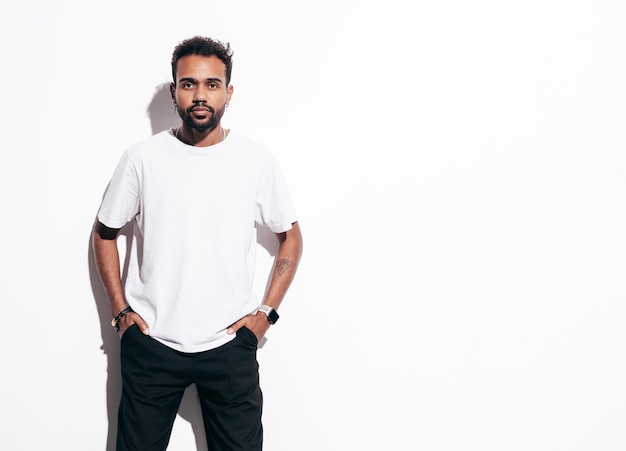 Guapo modelo hipster sonriente Hombre sexy sin afeitar vestido con camiseta de verano y ropa de jeans Hombre de moda posando junto a la pared blanca en el estudio en un día soleado Aislado