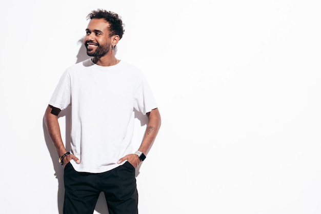 Guapo modelo hipster sonriente Hombre sexy sin afeitar vestido con camiseta de verano y ropa de jeans Hombre de moda posando junto a la pared blanca en el estudio en un día soleado Aislado