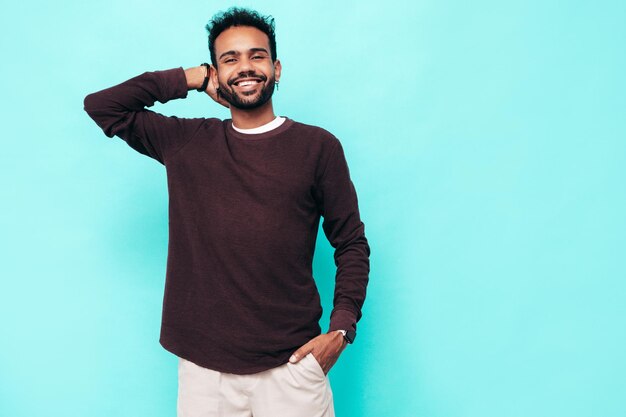 Guapo modelo hipster sonriente Hombre sexy sin afeitar vestido con suéter y ropa de jeans Hombre de moda posando junto a la pared azul en el estudio Mirando a la cámara