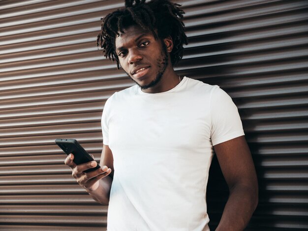 Guapo modelo hipsterHombre africano sin afeitar vestido con camiseta blanca de veranoHombre de moda con peinado de rastas Posando en la calleMirando la pantalla del teléfono inteligente usando aplicaciones de teléfonos celulares