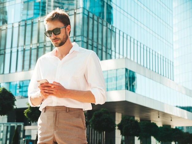 Guapo modelo lambersexual con estilo inconformista seguro Hombre moderno vestido con camisa blanca Hombre de moda posando en el fondo de la calle cerca de rascacielos Al aire libre al atardecer Usando aplicaciones de teléfonos inteligentes