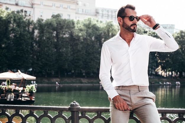 Guapo modelo lambersexual con estilo inconformista seguro Hombre moderno vestido con camisa blanca y pantalones Hombre de moda posando en la calle cerca del terraplén en la ciudad del parque de Europa al atardecer En gafas de sol