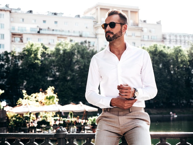 Guapo modelo lambersexual hipster con estilo seguro Hombre moderno sexy vestido con camisa blanca y pantalones Hombre de moda posando en la calle cerca del terraplén en la ciudad del parque de Europa al atardecer Con gafas de sol