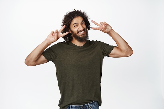 Foto gratuita guapo modelo masculino que muestra el gesto de paz vssign cerca de los ojos sonriendo y luciendo feliz de pie sobre fondo blanco copiar espacio