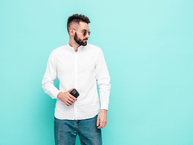 Guapo modelo seguro Hombre elegante sexy vestido con camisa y jeans Hombre hipster de moda posando cerca de la pared azul en el estudio Sosteniendo el teléfono inteligente Mirando la pantalla del teléfono celular Usando aplicaciones aisladas