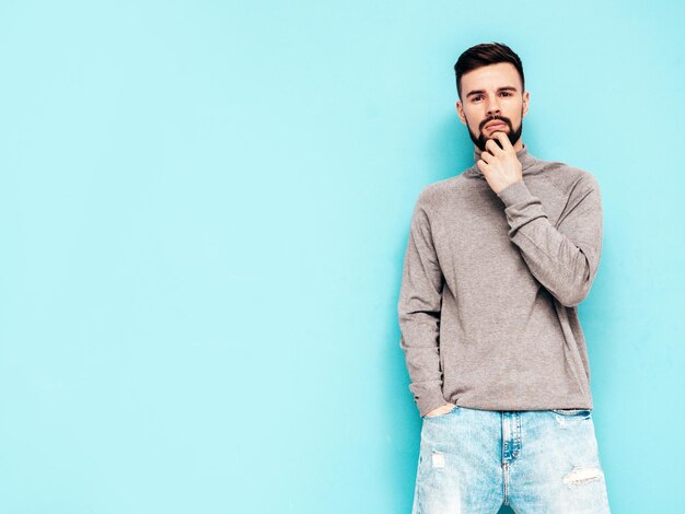 Guapo modelo sonriente Hombre elegante sexy vestido con suéter de cuello alto gris y jeans Hombre hipster de moda posando junto a la pared azul en el estudio Aislado