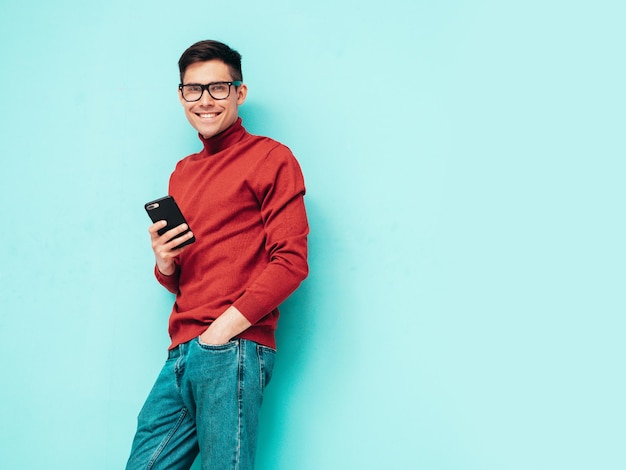 Guapo modelo sonriente Hombre elegante sexy vestido con suéter de cuello alto rojo y jeans Hombre hipster de moda posando junto a la pared azul en el estudio Sosteniendo teléfono inteligente Mirando la pantalla del teléfono celular Usando aplicaciones
