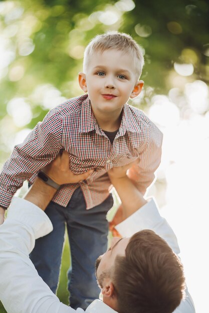 Guapo padre arrojando a un bebé contra el parque de verano Niño pequeño vestido con ropa elegante y papá pasando tiempo juntos sonriendo y divirtiéndose afuera en la naturaleza Concepto de crianza feliz