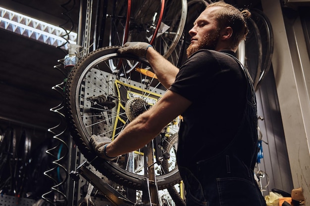 Foto gratuita un guapo pelirrojo con un mono de jeans, trabajando con una rueda de bicicleta en un taller de reparación. un trabajador quita el neumático de bicicleta en un taller.