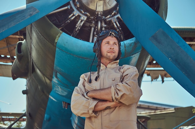 Guapo piloto en un equipo de vuelo completo de pie con los brazos cruzados cerca de un avión militar.