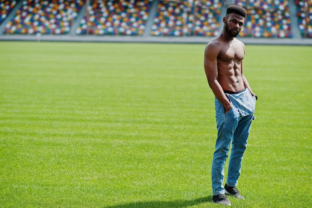 Guapo sexy afroamericano musculoso torso desnudo hombre en overoles de jeans posó en la hierba verde del campo de fútbol del estadio Retrato de hombre negro de moda