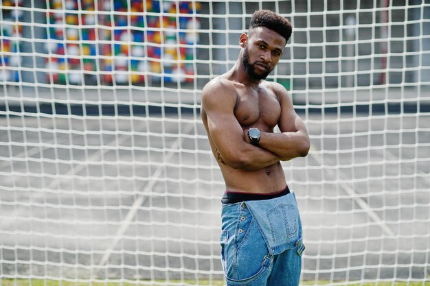 Guapo sexy afroamericano musculoso torso desnudo hombre en overoles de jeans posó en la hierba verde contra las puertas de fútbol del campo del estadio Retrato de hombre negro de moda