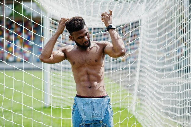 Guapo sexy afroamericano musculoso torso desnudo hombre en overoles de jeans posó en la hierba verde contra las puertas de fútbol del campo del estadio Retrato de hombre negro de moda