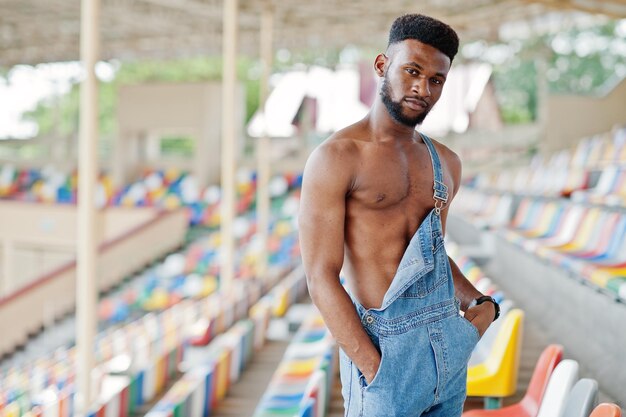 Guapo sexy afroamericano torso desnudo hombre en overoles de jeans planteados en sillas de colores en el estadio Retrato de hombre negro de moda