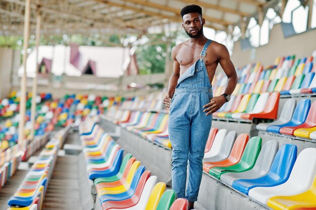 Guapo sexy afroamericano torso desnudo hombre en overoles de jeans planteados en sillas de colores en el estadio Retrato de hombre negro de moda