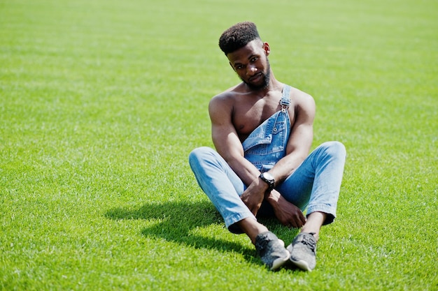Guapo sexy afroamericano torso desnudo hombre en overoles de jeans sentado en la hierba verde del campo de fútbol del estadio Retrato de hombre negro de moda