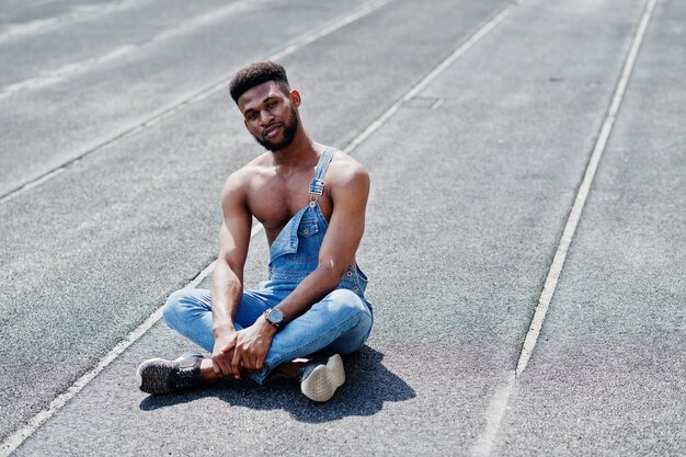 Guapo sexy afroamericano torso desnudo hombre en overoles de jeans sentado en el hipódromo del estadio Retrato de hombre negro de moda