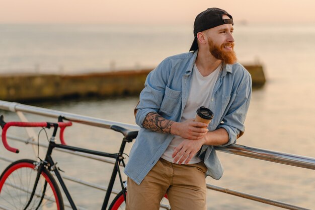 Guapo sonriente estilo hipster hombre con barba de jengibre vestido con camisa de mezclilla y gorra con bicicleta en el amanecer de la mañana junto al mar tomando café, viajero de estilo de vida activo y saludable