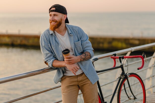 Guapo sonriente estilo hipster hombre con barba de jengibre vestido con camisa de mezclilla y gorra con bicicleta en el amanecer de la mañana junto al mar tomando café, viajero de estilo de vida activo y saludable