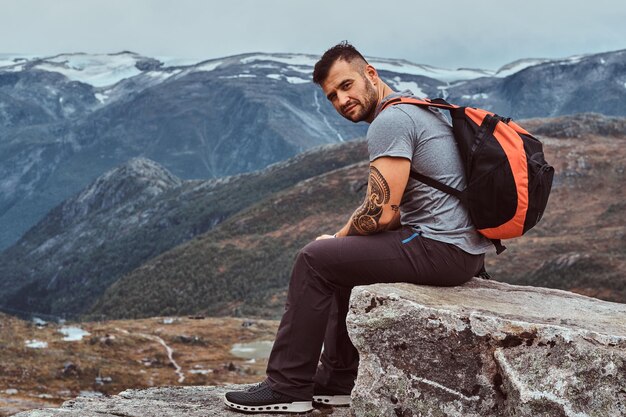 Guapo turista barbudo se sienta en una roca en el fondo de un hermoso paisaje montañoso en Noruega.