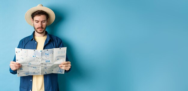 Guapo turista con sombrero de verano leyendo mapa en busca de turismo de vacaciones de pie en blue ba