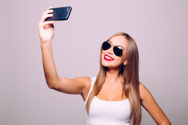 Guardando la memoria de su nuevo estilo. Retrato de hermosa mujer joven con gafas ajustando su sombrero mientras hace selfie y de pie contra la pared gris