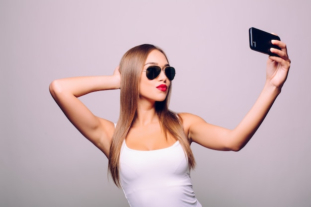 Guardando la memoria de su nuevo estilo. Retrato de hermosa mujer joven con gafas ajustando su sombrero mientras hace selfie y de pie contra la pared gris