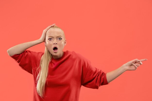 Guau. Hermoso retrato frontal de medio cuerpo femenino aislado sobre fondo de estudio coral. Joven mujer sorprendida emocional apuntando a la izquierda. Las emociones humanas, el concepto de expresión facial. Colores de moda