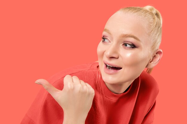 Guau. Hermoso retrato frontal de medio cuerpo femenino aislado sobre fondo de estudio de coral. Joven mujer sorprendida emocional apuntando a la izquierda. Las emociones humanas, el concepto de expresión facial. Colores de moda